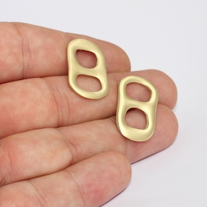 May include: Two matte gold-colored metal charms in the shape of a soda can tab. Each charm has two oval openings. The charms are displayed against a neutral background.