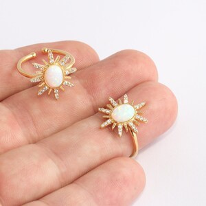 May include: Two gold-colored rings with a sunburst design. Each ring features a central oval-shaped white stone surrounded by small, clear crystals. The sunburst design extends outward from the central stone. One ring is open and adjustable.
