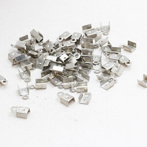 Puede incluir: Un conjunto de pequeños terminales de crimpado de metal plateado, dispersos sobre una superficie blanca. Cada terminal tiene un cuerpo rectangular con un lazo y una abertura en forma de U para sujetar a cordones o cintas. Para la fabricación de joyas.