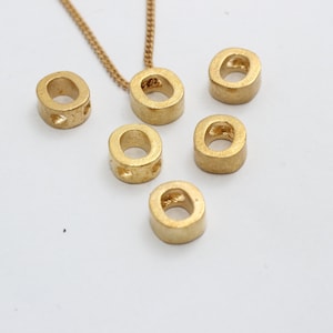 May include: A gold-colored necklace with a chain and a pendant in the shape of the letter "O". Several additional "O" shaped beads are scattered around the pendant. The beads have a matte finish.