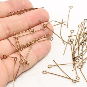 Puede incluir: Una colección de alfileres de ojo de color bronce, dispersos sobre una superficie blanca. Cada alfiler tiene un alambre recto con un extremo en bucle, ideal para la fabricación de joyas y manualidades. Los alfileres varían en longitud y son de metal.