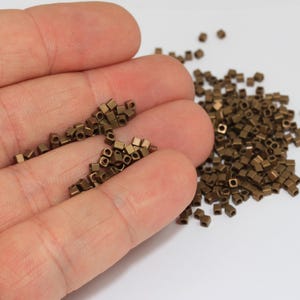 Pode incluir: Uma coleção de pequenas contas cúbicas de cor bronze metálico. Estas minúsculas contas quadradas apresentam um orifício central para enfiar e estão espalhadas por uma superfície, com parte das contas a descansar numa palma para escala.