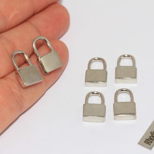 Puede incluir: Pequeños candados plateados con asas arqueadas. Dos candados se sostienen en una mano, mientras que otros cuatro están dispuestos sobre una superficie blanca. También hay una pequeña etiqueta rectangular con el texto "Rhodium Plated".