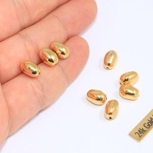 Może przedstawiać: Owalne koraliki platerowane złotem. Koraliki mają błyszczący złoty kolor i gładki, zaokrąglony kształt. Widoczna jest również mała prostokątna etykieta z napisem "24k Gold Plated".