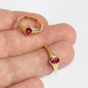 May include: Two gold rings, each featuring a teardrop-shaped, red gemstone. One ring is adjustable, the other a solid band. The rings are shown against a light background.