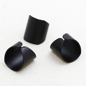 Puede incluir: Se muestran tres anillos negros y curvos sobre un fondo blanco. Los anillos tienen un acabado liso y mate y un diseño abierto único. Los anillos parecen estar hechos de un material sólido, posiblemente metal o plástico duradero.