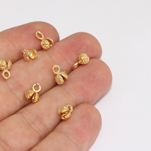 May include: Close-up of several small, gold-colored bead caps. Each cap has a textured, floral-like design and a small loop for attaching to jewelry. The caps are scattered against a white background, suggesting they are craft supplies.