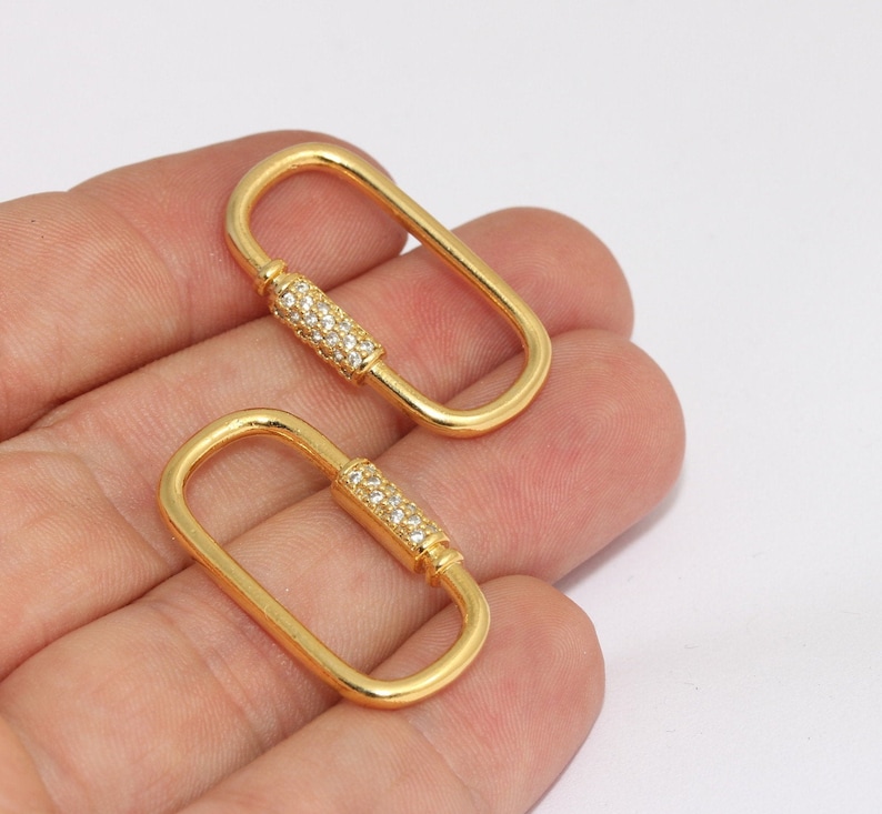 Op de afbeelding: Twee goudkleurige ovale karabijnhaaksluitingen. Elke sluiting heeft een centraal gedeelte ingelegd met kleine, heldere kristallen. De sluitingen worden in een hand gehouden tegen een witte achtergrond. Geschikt voor het maken van sieraden of knutselen.