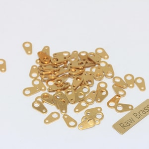 Pode incluir: Uma coleção de pequenos acessórios de latão dourado, incluindo formas ovais e retangulares com orifícios para fixação a outros materiais. Uma pequena etiqueta de latão com o texto "Raw Brass" também está presente.