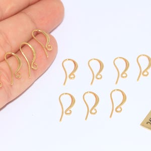 May include: Several gold-coloured earring hooks, some held in a hand, with a small rectangular tag that reads "24k Gold Plated". The hooks are in a variety of shapes, all with a curved design.