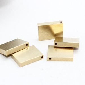 Puede incluir: Varios componentes metálicos pequeños, rectangulares y dorados, cada uno con un agujero perforado en un extremo, están dispuestos sobre una superficie blanca. Los componentes tienen un acabado liso y reflectante, probablemente para la fabricación de joyas.