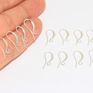 Puede incluir: Un conjunto de ganchos para pendientes de color plateado. Algunos ganchos se sostienen en una mano, mientras que otros están sobre una superficie blanca. Los ganchos tienen un diseño curvo con un extremo en bucle para sujetar los pendientes.