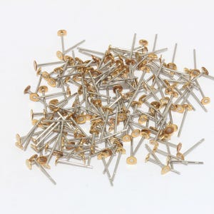 Puede incluir: Un montón de pequeños sujetadores metálicos de color plateado con cabezas circulares planas de color dorado. Los sujetadores tienen un poste largo, delgado y recto. Los objetos están dispersos sobre un fondo blanco.