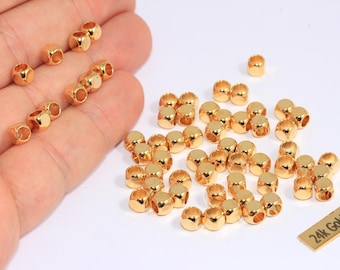 May include: A collection of small, gold-plated beads with a shiny finish. The beads are cylindrical with rounded edges and a hole running through the center. A small rectangular tag reads "24k Gold Plated".