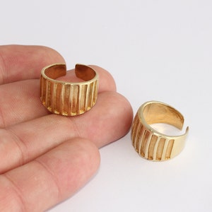 May include: Two gold-colored adjustable rings. Each ring has a thick band with vertical grooves. The rings are open at the top, allowing for size adjustment. The rings are displayed against a white background.