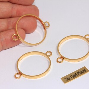 Puede incluir: Tres conectores de joyería circulares de metal dorado con un acabado liso. Cada anillo cuenta con dos pequeñas anillas en los lados opuestos para su fijación. Una pequeña etiqueta rectangular incluye el texto 24k Gold Plated.