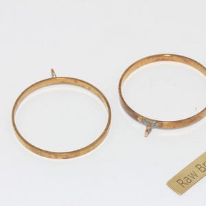 Puede incluir: Dos marcos circulares de latón en bruto con pequeños bucles para colgar. También hay una pequeña etiqueta rectangular de latón con las palabras "Raw Brass". Los marcos son de color dorado cálido y están sobre una superficie blanca.