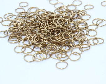 May include: A pile of small, gold-colored jump rings on a white background. The rings are open and ready for use in jewelry making or other crafts. The rings are made of metal.