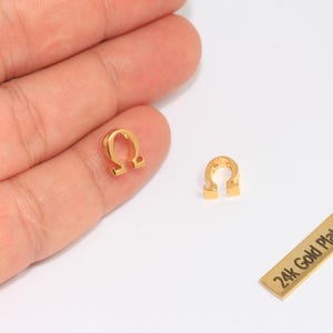 Pode incluir: Close-up de três achados de joalheria banhados a ouro. Dois têm a forma da letra grega Ômega, e o terceiro é uma barra retangular com o texto "24k Gold Plated". Os achados estão em uma superfície branca.