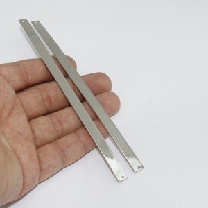 Pode incluir: Duas barras de metal retangulares prateadas com bordas arredondadas e um pequeno orifício em uma extremidade. As barras são seguradas em uma mão contra um fundo branco. Provavelmente achados de joias ou componentes de artesanato.