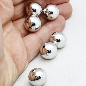 Könnte beinhalten: Eine Sammlung glänzender, silberfarbener, kugelförmiger Perlen. Die Perlen sind glatt und reflektierend, mit einem kleinen Loch oben. Die Perlen werden in der Hand einer Person gezeigt.