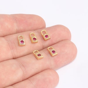 May include: Close-up of several small, gold-colored padlock charms. Each charm has a rectangular body with a rounded top and a small, round, red gemstone set in the center. The charms are displayed against a light background.
