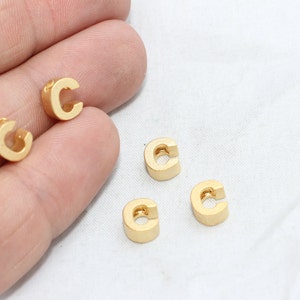 May include: Four small, gold-colored letter "C" charms are displayed. The charms are made of a solid, metallic material and have a rounded shape. The charms are scattered on a white surface.