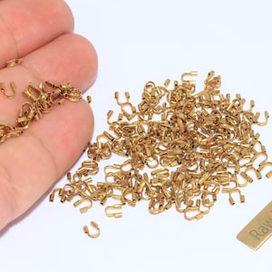 Op de afbeelding: Een verzameling kleine, goudkleurige messing fournituren, waaronder U-vormige connectoren en andere decoratieve elementen. De fournituren zijn verspreid over een wit oppervlak, met een klein rechthoekig label met de tekst "Raw Brass".