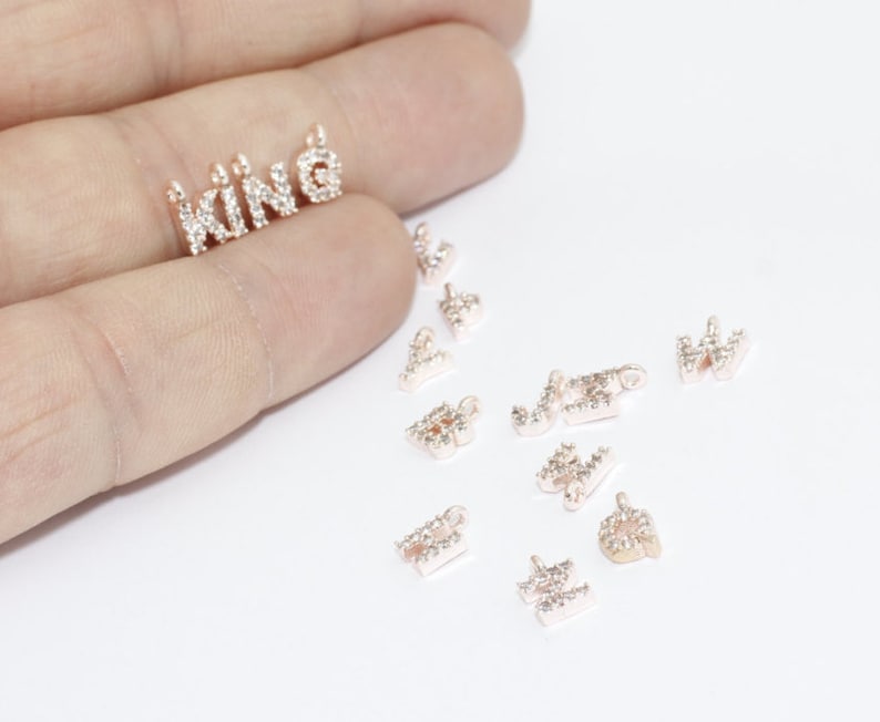 May include: Rose gold-toned metal charms, including the word "KING" and individual letters, are encrusted with small, clear crystals. The charms are designed for jewelry making or crafting. The letters are small and delicate.