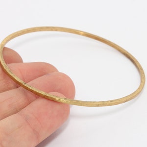 Op de afbeelding: Een gehamerde messing armband. De dunne, ronde armband heeft een getextureerd oppervlak en een warme, gouden kleur. De armband wordt tegen een witte achtergrond gehouden.