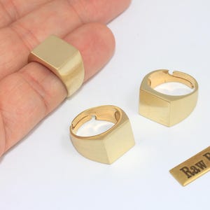 Könnte beinhalten: Goldfarbene Messingringe mit quadratischem Design. Ein Ring befindet sich an einem Finger, zwei weitere werden separat angezeigt. Ein kleines Messingrechteck mit dem Text "Raw Brass" ist ebenfalls vorhanden.