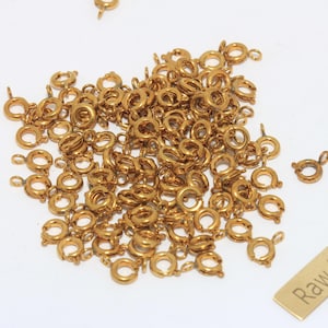 Puede incluir: Un montón de pequeños cierres de resorte dorados para joyería. Los cierres son redondos con un pequeño lazo y una palanca. También hay una pequeña etiqueta de latón con el texto "Raw Brass".