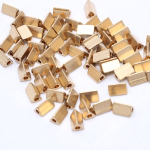 May include: A collection of small, gold-colored metal beads. The beads have a rectangular shape with a triangular end and a hole for stringing. They are scattered on a white surface.