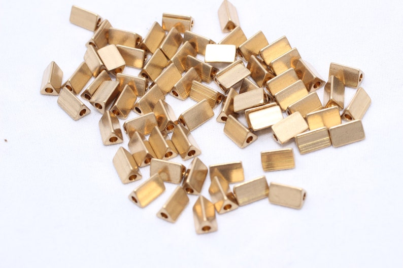 May include: A collection of small, gold-colored metal beads. The beads have a rectangular shape with a triangular end and a hole for stringing. They are scattered on a white surface.