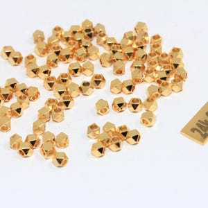 May include: A collection of small, gold-plated, faceted beads scattered on a white surface. A small rectangular tag with the text "24k Gold Plated" is also present. The beads have a geometric shape and are designed for jewelry making.
