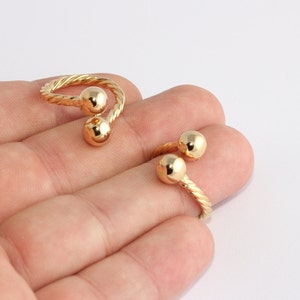 May include: Two gold-colored, adjustable rings with a twisted band design. Each ring features two spherical accents. The rings are displayed on a light background.
