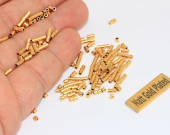 May include: A selection of small, gold-coloured metal tubes and cylinders, some held in a hand, scattered on a white surface. A rectangular tag with the text "Matt Gold Plated" is also present. These appear to be jewellery findings or small hardware components.