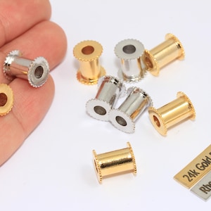 Puede incluir: Un surtido de hallazgos de joyería dorados y plateados, que incluyen componentes cilíndricos con un borde texturizado. Dos pequeñas etiquetas rectangulares dicen "24k Gold Plated" y "Rhodium Plated". Los artículos se muestran sobre una superficie blanca.