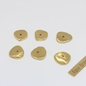 May include: Six irregular-shaped, gold-colored brass beads with a small hole in the center. A small brass tag with the text "Raw Brass" is also present. The beads have a smooth, polished finish.