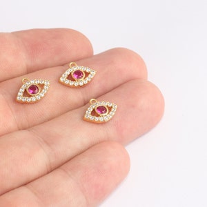 Peut inclure: Trois breloques dorées en forme d'œil. Chaque breloque présente une pierre précieuse rose centrale entourée de petits cristaux transparents. Les breloques sont conçues avec une petite boucle en haut pour l'attache, pour la fabrication de bijoux.