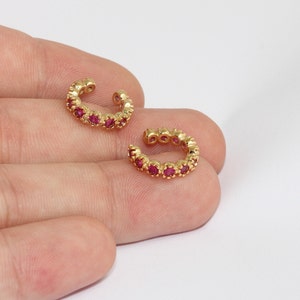 May include: Two gold-colored ear cuffs, each adorned with a row of small, vibrant red gemstones. The ear cuffs are crescent-shaped, designed to wrap around the ear's cartilage. The gemstones are evenly spaced, creating a continuous line of color.