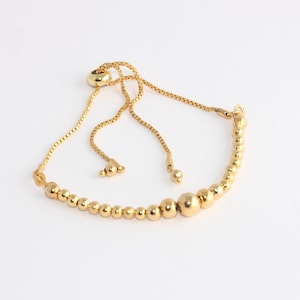 May include: Gold-colored adjustable bracelet featuring a series of small, round beads that gradually increase in size towards the center. The bracelet has a delicate chain and a sliding bead closure for adjustment.