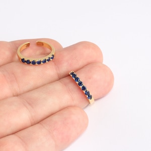 May include: Two gold-colored rings with a row of small, dark blue gemstones. One ring is an adjustable toe ring, and the other is a thin band. The rings are displayed on a light background.