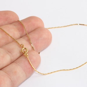 May include: A delicate gold-colored necklace with a fine chain. The necklace features a small clasp and is displayed against a white background. The chain is thin and appears to be made of small, rectangular links.