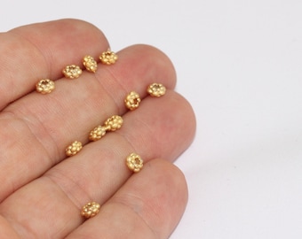 May include: Close-up of small, gold-colored beads with a textured, floral-like design. The beads are scattered on a light background, suggesting their use in jewelry making or other crafts.