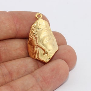 May include: A gold-coloured pendant with a face design. The pendant has a textured surface and a loop at the top for hanging. The face features closed eyes and a defined nose and mouth. The pendant is held in a hand against a white background.