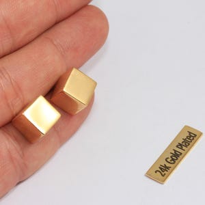 May include: Two gold-plated cube-shaped earrings and a small rectangular tag with the text "24k Gold Plated". The earrings are held in a hand.