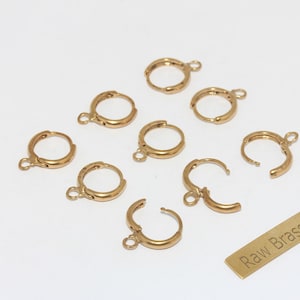 May include: A collection of gold-colored leverback earring findings. The earrings are in various states of closure, with some open and some closed. A small rectangular tag with the text "Raw Brass" is also present.