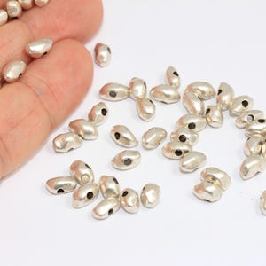 May include: Silver-coloured, oval-shaped beads with a matte finish and a small hole on each end. The beads are scattered on a white surface, with some held in a hand. These beads are suitable for jewellery making and crafting projects.
