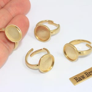 May include: Several adjustable brass rings with oval settings are displayed. One ring is on a hand. A small brass tag with the text "Raw Brass" is also present. These rings are suitable for jewelry making.